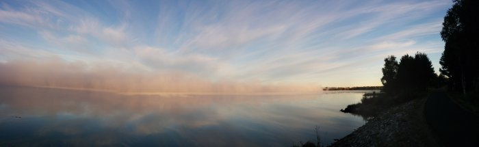 Morgondimma lägger sig tätt över stilla Kalix älv med träd och väg vid sidan, tidig morgon.