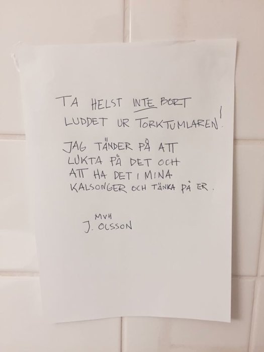 Ett meddelande upptejpat på en vit kakelvägg med en handskriven text som ber om att inte ta bort luddet ur torktumlaren.