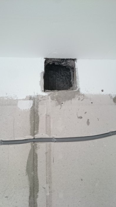 Självdragshål i en vitmålad vägg över en flexibel elkabel, omringat av ojämnt spackel och sotiga kanter.
