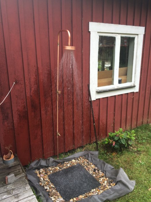 Utomhusdusch med kopparhuvud mot rödmålad vägg vid ett hus, flankerad av gröna växter.