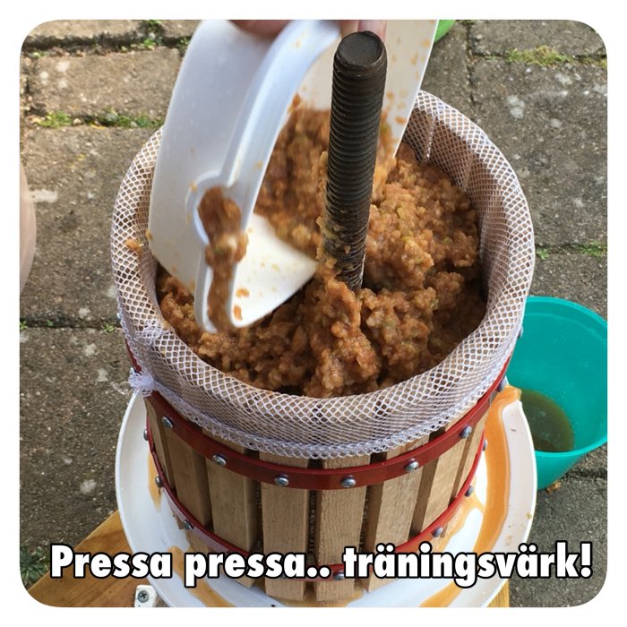 Äppelmos pressas i en fruktpress över en hink, med texten "Pressa pressa... träningsvärk!