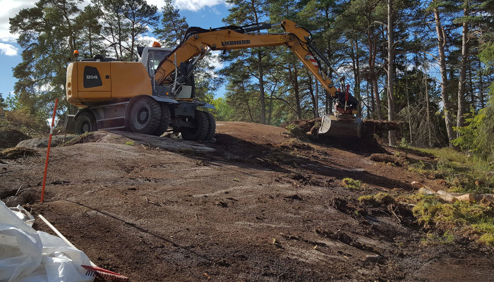 Grävmaskin som skalar av berg på en byggarbetsplats med skog omkring.