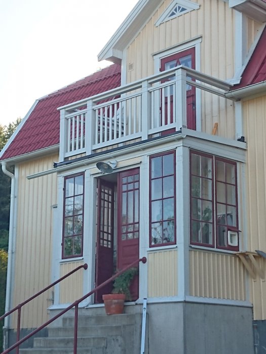 Nästan färdigbyggd balkong på ett gult trähus med röda fönsterkarmar och en matchande röd takpannor.