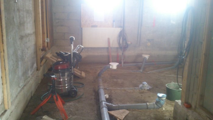 Ofärdigt källargolv med synliga rörinstallationer och byggmaterial inför gjutning, dammsugare och verktyg syns.