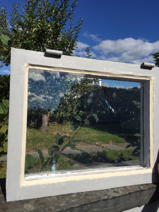 Lagat källarfönster med ny plexiglasskiva, omgiven av vitmålat fönsterkarm mot trädgård.