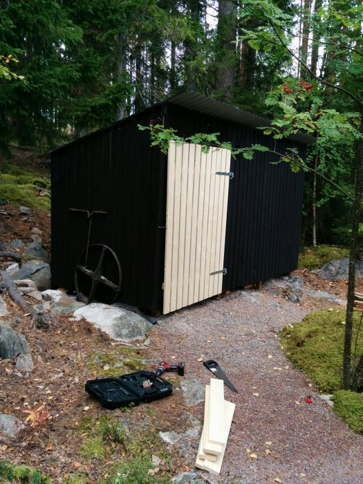 Nybyggd obehandlad träddörr på en svart vedbod, verktyg på marken framför.