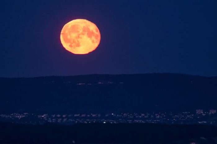 Fullmåne i orange nyanser stiger över silhuetten av en bergsrygg med stadslys nertill.