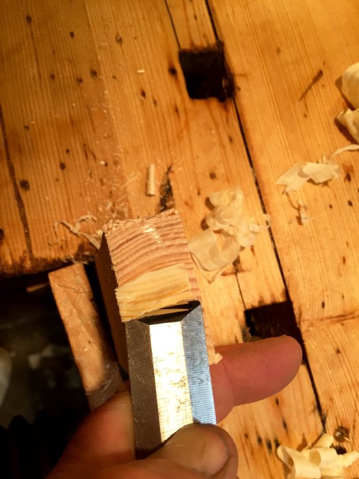 Hand som håller ett stämjärn med nyvässad egg mot en träbit med hyvelspån i en verkstadsmiljö.