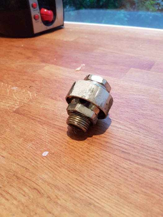 Metallisk anslutningsdel till vattenutkastare med tecken på läckage, ligger på ett träbord.