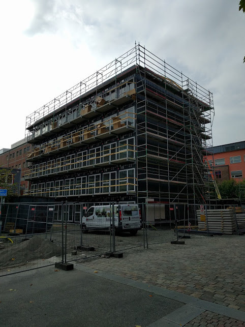 Byggnad under renovering med ställningar runt omkring, omgivet av byggmaterial och säkerhetsstaket.