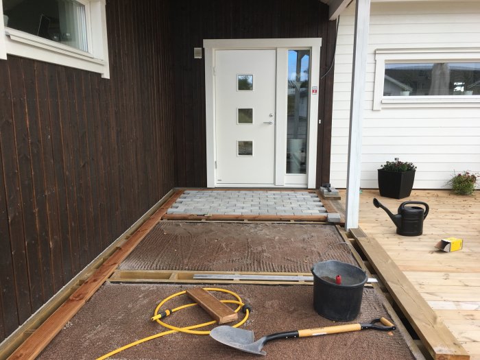 Pågående arbete med entrérenovering, halvlagd marksten framför vit dörr, trallram till höger och byggverktyg på sandig yta.