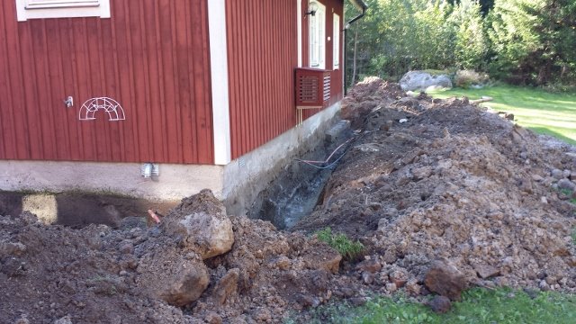 Grävd grund vid röd husvägg med synligt dräneringsrör, grushög och utsatt grundmur.