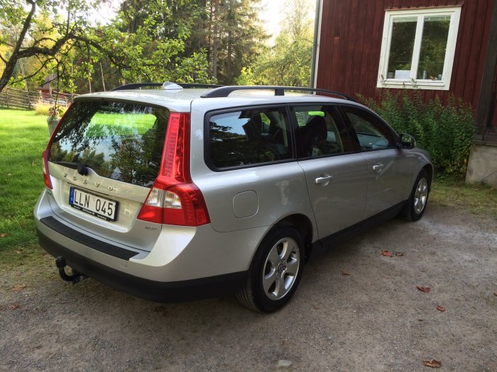 Silverfärgad Volvo V50 parkerad framför röd stuga med grönskande omgivning.