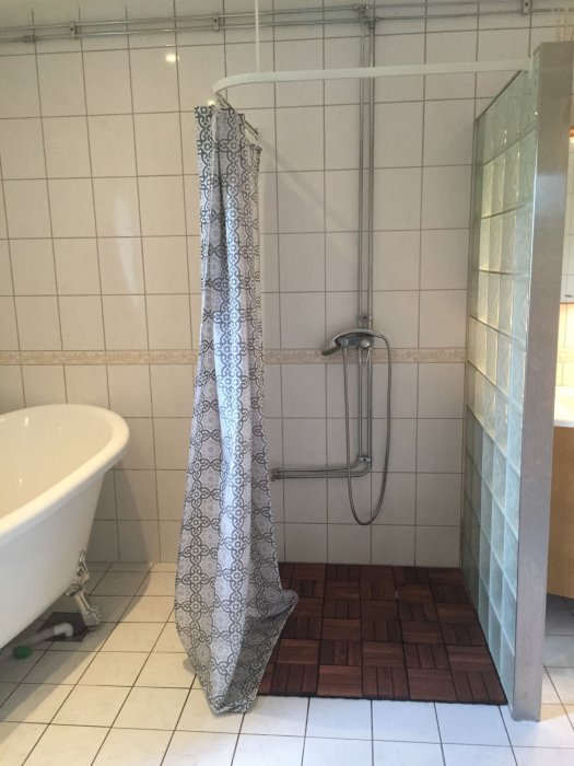 Badrum med vit kakelvägg, utanpåliggande rör och befintlig dusch upphängd vid en duschstång med mönstrad duschdraperi.