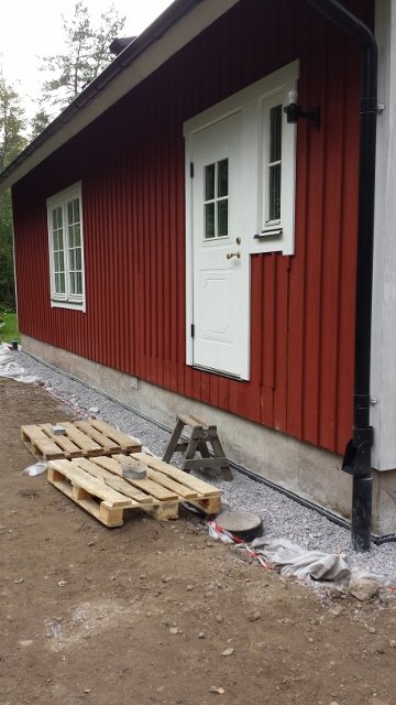 Rödmålad husvägg med vit ytterdörr och en oavslutad veranda under konstruktion, omgiven av grus och byggmaterial.