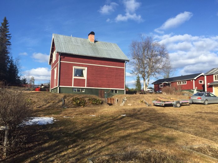 Rödmålad stuga utan tak över källaringång, byggmaterial på släpvagn, vårlandskap.