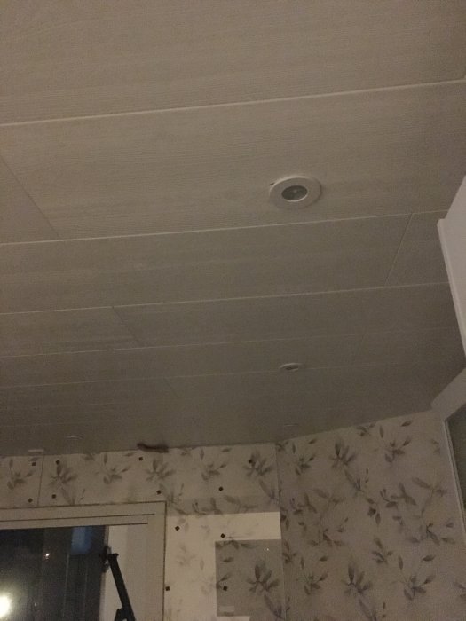 Nypanelad vägg och tak med spotlights, oavslutat fönsterfoder och blommigt tapet.