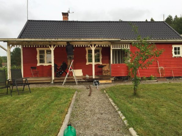 Person på stege framför rött trähus under renovering, med gräsmatta och stig i förgrunden.