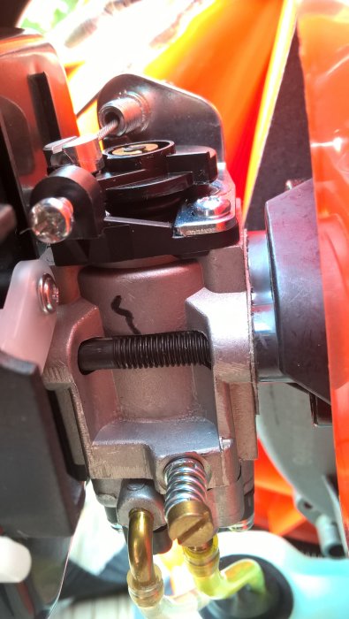 Närbild på VidaXL-maskinens förgasare med synliga justeringsskruvar och fjädrar mot orange bakgrund.
