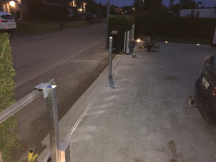 Kvällsbild på trottoar med staket i byggnad, parkerade bilar, utebelysning och trädgårdsarbetsredskap i förortsmiljö.