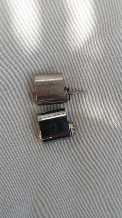 Två låscylindrar på tygunderlag varav en med en längre plåtbit för dörrlåsning.
