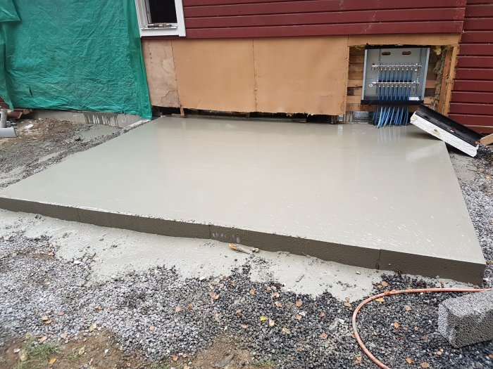 Nyligen gjuten självkompakterande betongplatta vid husgrund med grus och elskåp i bakgrunden.