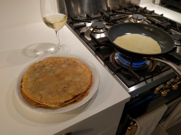 Pannkakstårta på tallrik bredvid spis med stekpanna och en pannkaka som tillagas, ett glas vitt vin på bänk.