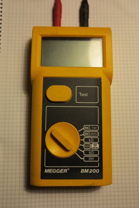 Gul och svart Megger BM200 isolationsprovare med testknapp och inställningsratt mot ett rutigt underlag.