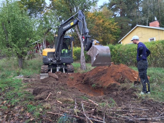 Grävmaskin och person som arbetar med att gräva upp VA-ledning i trädgård med hus i bakgrunden.