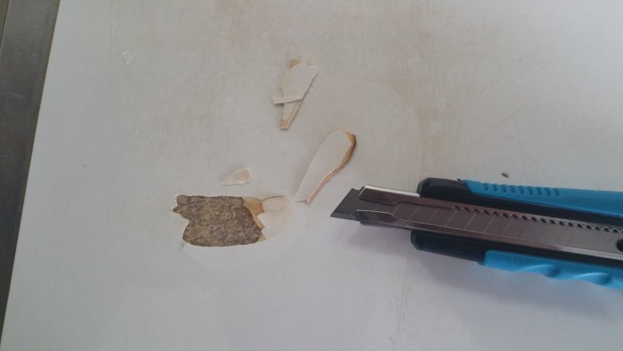 Skadad laminatbänkskiva med lossade bitar och en skrapa för reparation.