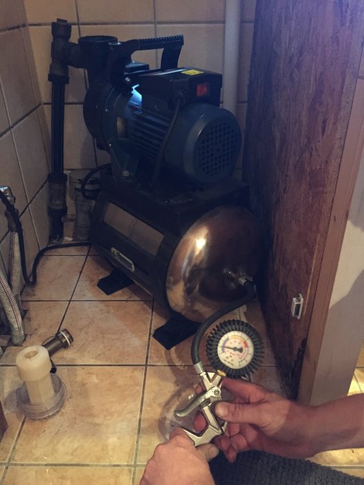 Vattenpump och trycktank i en husgrund med synlig tryckmätare och avskruvad vattenkoppling.