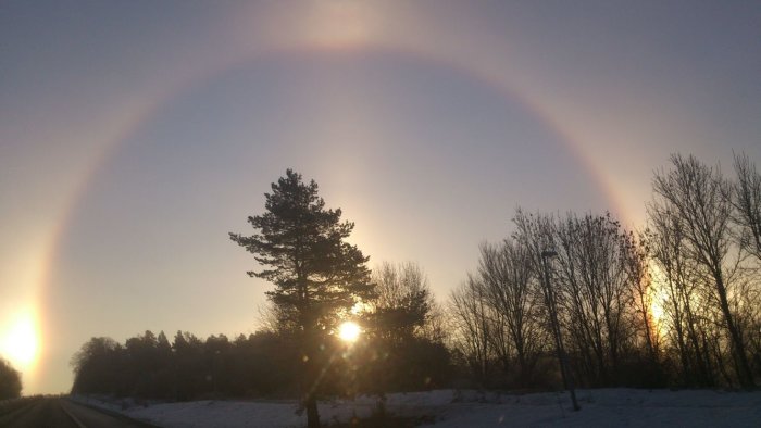 Halofenomen med en stor cirkel runt solen och två bisolar synliga från en snöig väg.