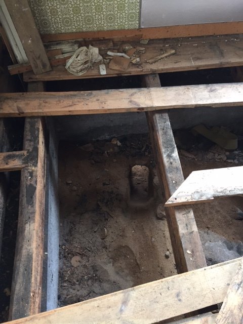 Uppgrävt golv i tvättstuga med synliga träbalkar och en grävd grop för installation av avlopp.