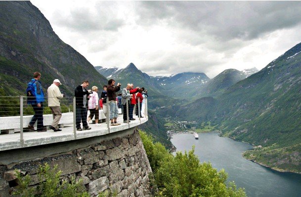 Besökare betraktar utsikten från en utkiksplattform på en klippa ovanför en fjord i Norge, räcket ser smalt ut.