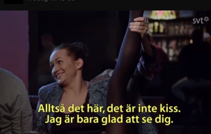 Kvinna ler, lyfter arm, två personer, bar-miljö, svensk text: "inte kiss, bara glad".