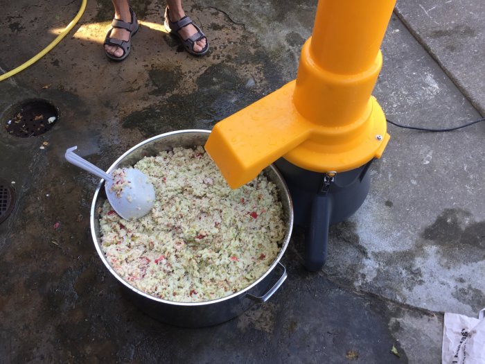 Krossade äpplen i en stor metallgryta bredvid en gul och svart äppelpress ute på en stenlagd gård.