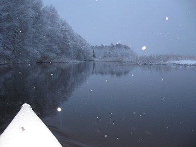 Vit kajakspets som sticker fram mot en mörk vinterflod omgiven av snöklädda träd och fallande snöflingor.