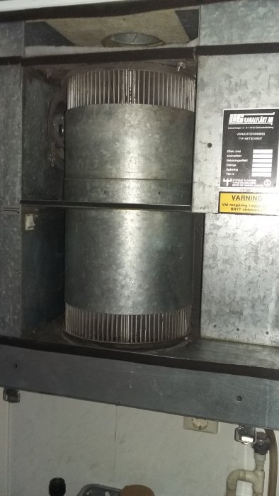 Äldre kanalfläkt i ett ventilationsutrymme med varningsskylt och synlig kabeldragning.