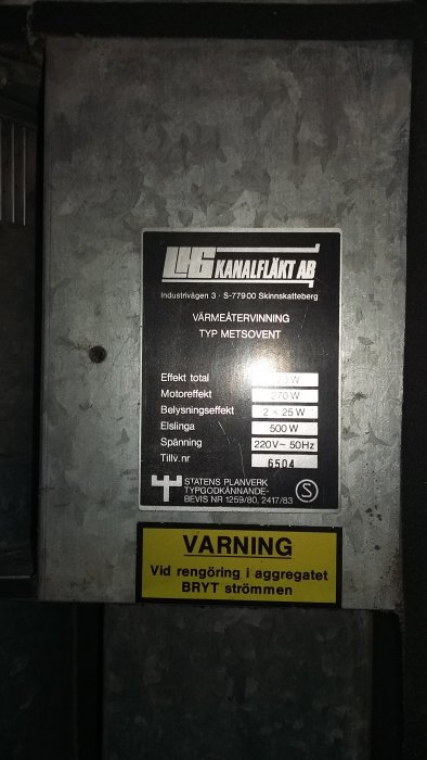 Äldre kanalfläkt med teknisk etikett och varningslapp i ett ventilationssystem från 1989.