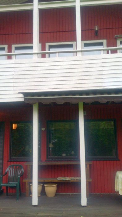 Fasad av hus med rött träpanel och vit balkong, gröna fönster och vit pelare, trädgårdsmöbler på verandan.