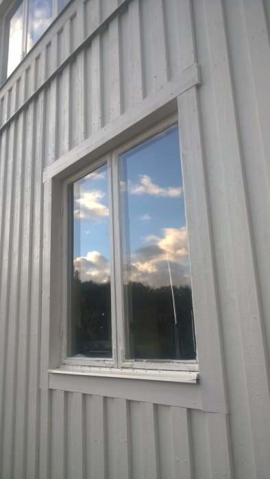 Fönster med vit karm monterat på ett hus med vertikal vit panel, reflekterande himmel och moln.