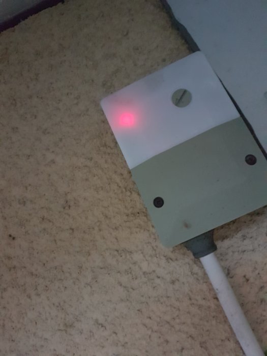 Väggmonterad elanordning med röd lampa och vred, sladd ansluten, mot texturerad bakgrund.