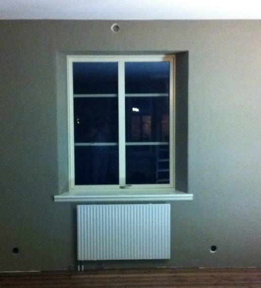 Nyinstallerat fönster ovanför en radiator i ett rum under renovering, med väggar gipsade i grått.