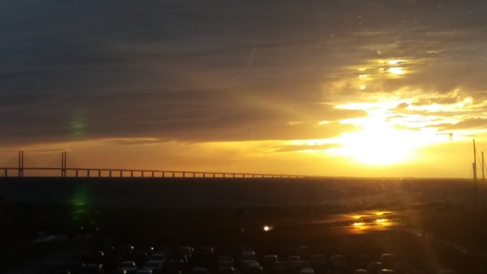 Solnedgång över en bro i Sverige med reflektioner och parkerade bilar i förgrunden.