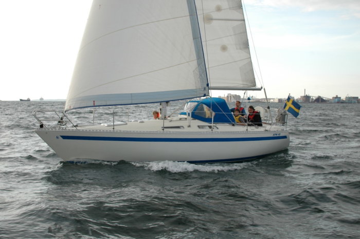 Segelbåt med svensk flagga navigerar på vågigt vatten, flera personer ombord.