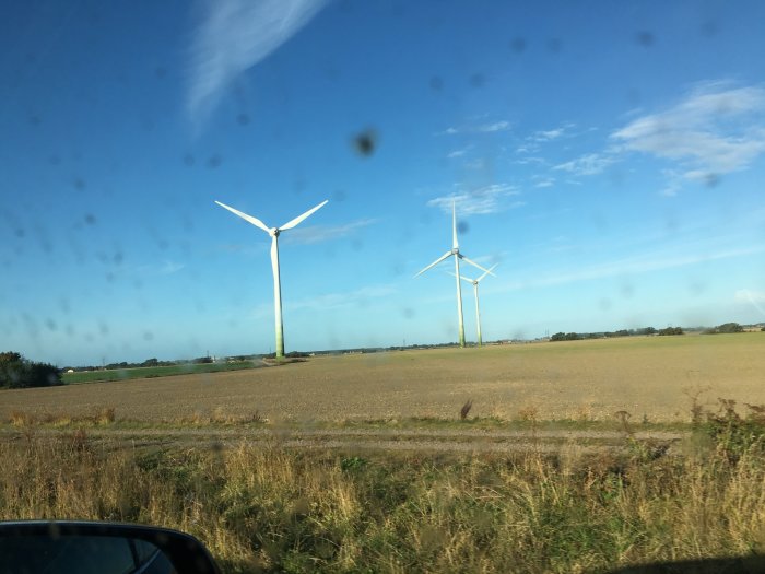 Vy över ett öppet landskap med två vindkraftverk mot en blå himmel, sedd genom en smutsig bilruta.