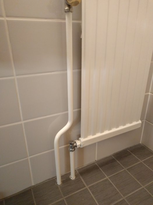 Nymålade vita elementrör och radiator mot kaklad vägg över mörkgrått klinkergolv.