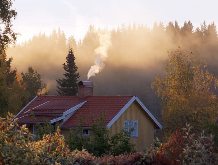 Gul husfasad med röda takpannor, skorsten med rök och höstträd i dimmigt morgonljus.