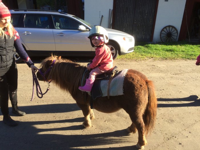 Ett barn i rosa hjälm och stövlar rider på en ponny hållen av en kvinna utomhus.