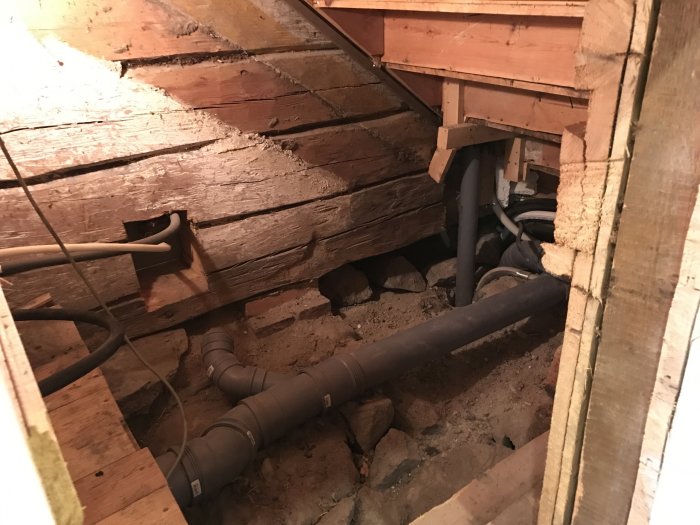 Öppnat utrymme under en trappa med synliga träbjälkar och rörledningar, och spår av borttagen isolering.